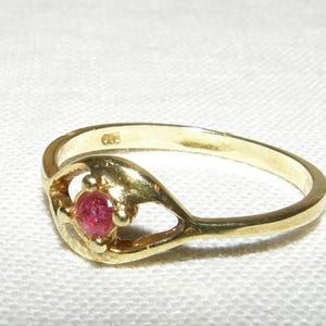 Puede incluir: Un anillo de oro con una pequeña gema roja y redonda en el centro. El anillo tiene un diseño abierto y único con un patrón arremolinado alrededor de la gema. El anillo está sobre un fondo blanco.