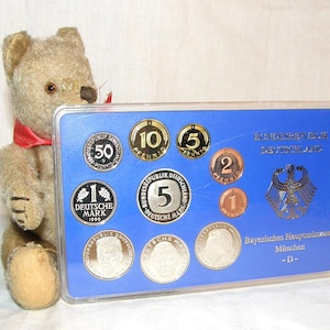 Peut inclure: Un ours en peluche vintage avec un ruban rouge est assis à côté d'un présentoir bleu de pièces allemandes. Le présentoir présente diverses pièces avec des dénominations telles que 50 Pfennig, 1 Deutsche Mark et 5 Pfennig. Le texte sur le présentoir indique "Bundesrepublik Deutschland" et "Bayerisches Hauptmünzamt München".