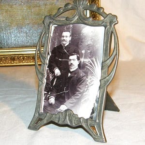 Pode incluir: Moldura de fotos de metal antiga com um retrato a preto e branco de dois homens. A moldura tem um design Art Nouveau ornamentado e um suporte embutido. A moldura é de cor cinza suave.