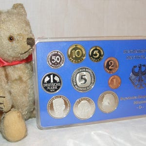 Può includere: Un orsacchiotto beige con un nastro rosso accanto a un espositore blu contenente monete tedesche. L'espositore presenta varie monete, tra cui 50 Pfennig e 1 Deutsche Mark. L'espositore ha testo in tedesco e un'aquila tedesca.