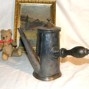 Op de afbeelding: Antieke zilverkleurige metalen koffiepot met een lange tuit en een donkerhouten handvat. De pot heeft een deksel en staat op een ronde voet. Een kleine teddybeer met een rode strik en een ingelijst schilderij staan op de achtergrond.
