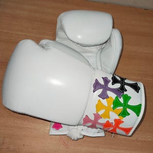 Könnte beinhalten: Weiße Boxhandschuhe mit bunten Kreuzmustern am Handgelenk. Die Kreuze sind rosa, orange, gelb, grün, lila und schwarz. Die Handschuhe sind aus Leder und haben eine gepolsterte Konstruktion.