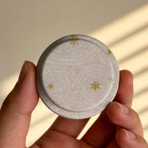 May include: A round, off-white lid with a decorative pattern of interlocking shapes and gold starburst designs. The lid is held in a hand, showcasing its detailed surface and color palette.
