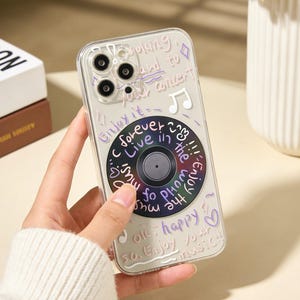 Puede incluir: Funda transparente para teléfono con diseño de disco de vinilo. La funda tiene texto escrito a mano y notas musicales. El diseño del disco de vinilo está en el centro. El texto incluye las palabras "Enjoy it", "forever" y "Enjoy your music".