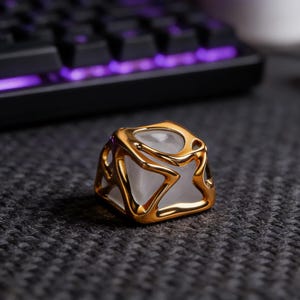 May include: A gold-colored, geometric keycap with a translucent center. The keycap is square-shaped with an open, artistic design. It sits on a dark gray surface, with a keyboard and a white mug in the background.