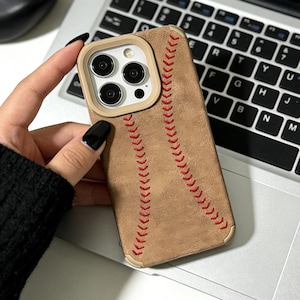May include: A tan phone case with red stitching, resembling a baseball. The case has a beige border around the camera lenses. The phone case is held over a laptop keyboard.