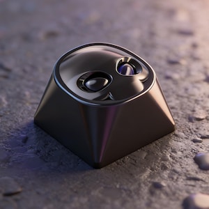 May include: A black, metallic keycap with a skull design. The keycap has a smooth, reflective surface and features eye and mouth details. The keycap is set against a textured, gray surface with water droplets.