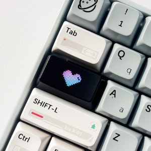 May include: Close-up of a mechanical keyboard with white and black keycaps. The black keycap features a pixelated heart design in shades of pink and blue. Other keys include symbols and letters, such as "Tab", "Shift-L", and "Ctrl".