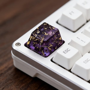 以下が含まれることがあります： カスタムキーボードのキーキャップのクローズアップ。キーキャップは、金色のフレークが埋め込まれた半透明の紫色です。他の白いキーが見える、白とシルバーのキーボードに取り付けられています。