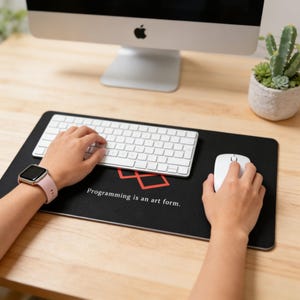 May include: A rectangular black desk mat featuring a red geometric logo and the white text "Programming is an art form." The mat is positioned under a white wireless keyboard and a white computer mouse on a light-colored wooden surface.