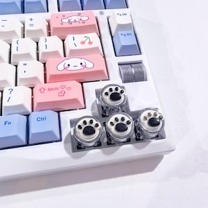 May include: A white mechanical keyboard with custom keycaps. Some keycaps are pink with cartoon characters and cherries. Others are white and blue. Four clear keycaps with black paw prints are in the foreground.