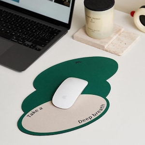 May include: A green and beige mousepad with the words "Take a deep breath" printed on it. A white computer mouse sits on the mousepad, next to a laptop and a candle.