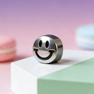 May include: A silver-colored smiley face charm with black details. The charm is round and has a shiny, reflective surface. The charm is displayed on a white and green geometric surface against a pink and purple background.