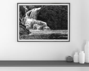 May include: Black and white framed photograph of a waterfall cascading over rocks into a river. The image captures the power of nature with the water's movement and the surrounding forest.