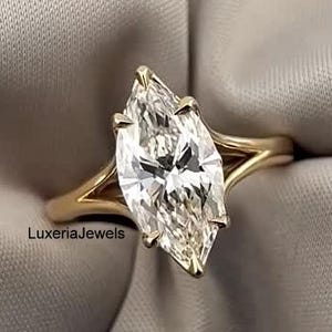 Op de afbeelding: Een verlovingsring met een marquise-geslepen diamant en een gouden band. De diamant is gezet in een vier-klauwzetting en de band heeft een split-schachtontwerp. De ring wordt getoond op een grijze stof.