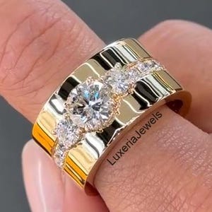 Op de afbeelding: Een brede, goudkleurige ring met een grote, heldere edelsteen in het midden en kleinere, heldere edelstenen aan weerszijden. De ring zit om een vinger en het oppervlak is reflecterend. De tekst "LuxeriaJewels" is zichtbaar.