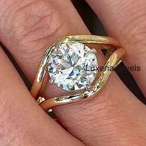 Pode incluir: Um anel de ouro com uma gema grande, redonda e transparente. A banda do anel curva-se em torno da gema, criando um design único e moderno. O anel é mostrado num dedo.