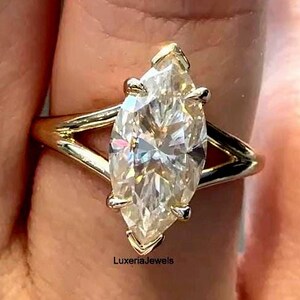 Pode incluir: Anel de diamante com corte marquise e banda dourada. O diamante é cravejado com quatro garras e tem um brilho brilhante. O anel é uma joia fina.