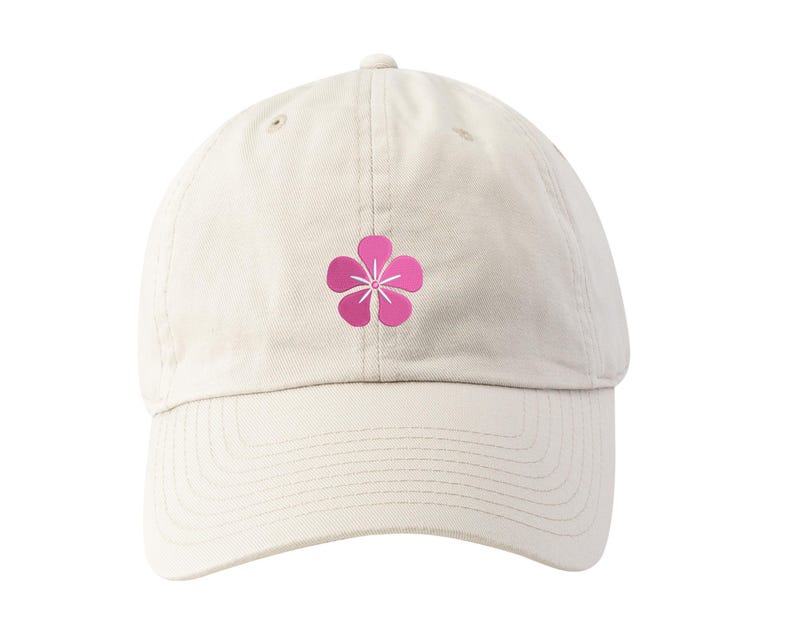 May include: A cream-colored baseball cap featuring a pink flower graphic on the front. The cap has a curved brim and an adjustable strap at the back. The flower design is a simple, five-petal blossom with a white center.