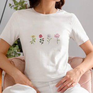 May include: White t-shirt featuring four embroidered flowers: a yellow chrysanthemum, a red rose, a blue daisy, and a pink lotus. The flowers are arranged horizontally across the chest. The shirt has short sleeves and a crew neck.