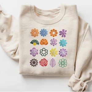 May include: A cream-colored sweatshirt featuring a grid of colorful flower designs. The flowers are in various colors, including orange, blue, purple, and green. The sweatshirt has a crew neck and long sleeves.
