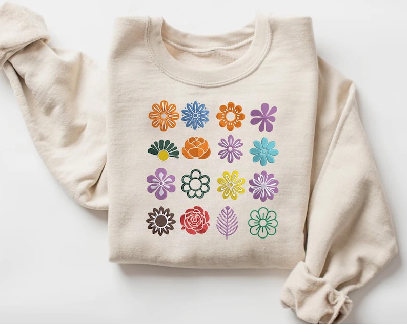 May include: A cream-colored sweatshirt featuring a grid of colorful flower designs. The flowers are in various colors, including orange, blue, purple, and green. The sweatshirt has a crew neck and long sleeves.