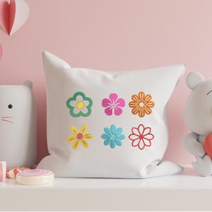 May include: A white square pillow with six colorful embroidered flowers. The flowers are in shades of green, pink, orange, yellow, blue, and red. The pillow is part of a decorative display with a cat-shaped light and a teddy bear.