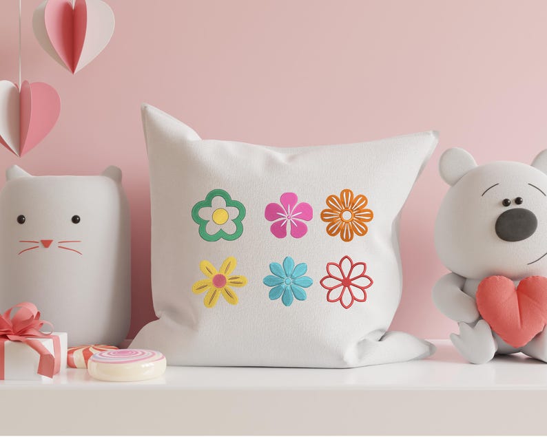 May include: A white square pillow with six colorful embroidered flowers. The flowers are in shades of green, pink, orange, yellow, blue, and red. The pillow is part of a decorative display with a cat-shaped light and a teddy bear.