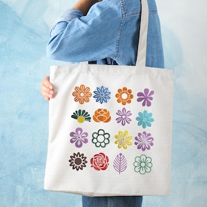 May include: A white tote bag featuring a colorful floral pattern. The design includes various flower illustrations in shades of orange, blue, purple, green, and yellow. The bag is held by a person wearing a denim jacket.