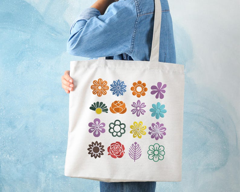 May include: A white tote bag featuring a colorful floral pattern. The design includes various flower illustrations in shades of orange, blue, purple, green, and yellow. The bag is held by a person wearing a denim jacket.