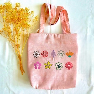 May include: A pink tote bag with a floral design. The bag has a rectangular shape with two handles. The floral design includes various colorful flower and leaf illustrations. A bundle of yellow dried flowers is placed next to the bag.