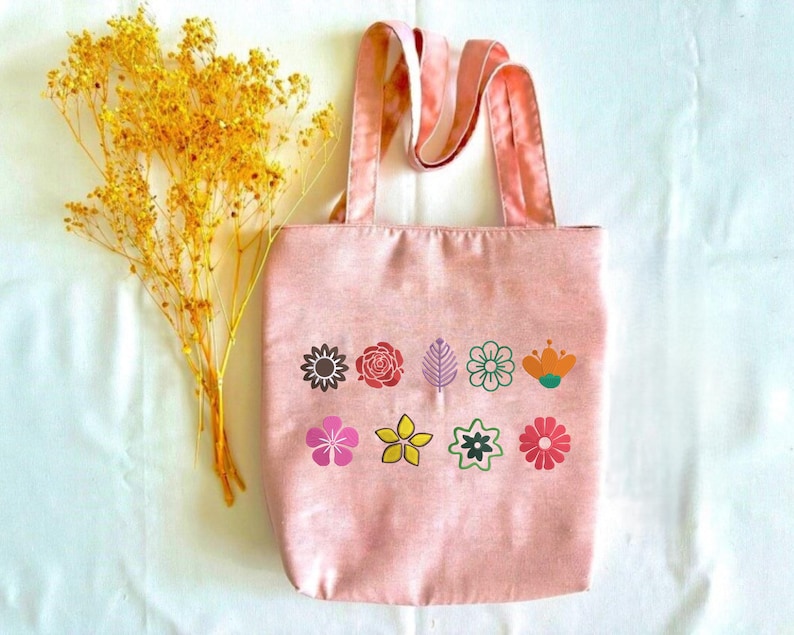 May include: A pink tote bag with a floral design. The bag has a rectangular shape with two handles. The floral design includes various colorful flower and leaf illustrations. A bundle of yellow dried flowers is placed next to the bag.