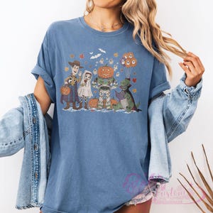 K&ouml;nnte beinhalten: Blaues T-Shirt mit einem Halloween-Motiv. Das Design zeigt Zeichentrickfiguren in Kost&uuml;men, darunter Woody, Buzz Lightyear und ein Dinosaurier, mit K&uuml;rbissen und Geistern. Das T-Shirt hat kurze &Auml;rmel.