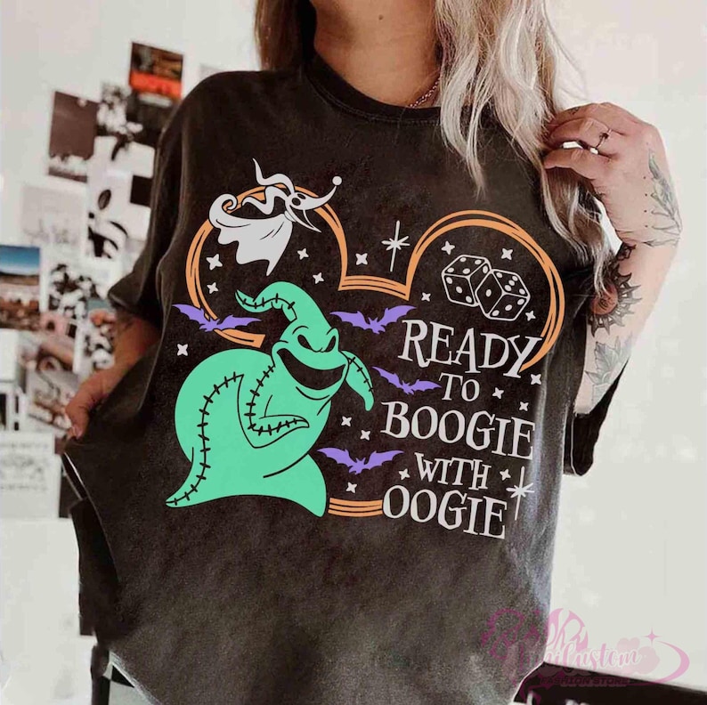 May include: A charcoal gray t-shirt featuring a graphic of a green Oogie Boogie character, a white Zero ghost dog, two dice, and purple bats. The text reads READY TO BOOGIE WITH OOGIE, arranged within a large orange outline of a mouse-ear shape.