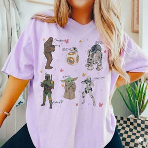 May include: A lilac t-shirt with illustrations of Star Wars characters: Chewbacca, BB-8, R2-D2, Boba Fett, Grogu, and a Stormtrooper. The characters' names are written above them in a handwritten style.