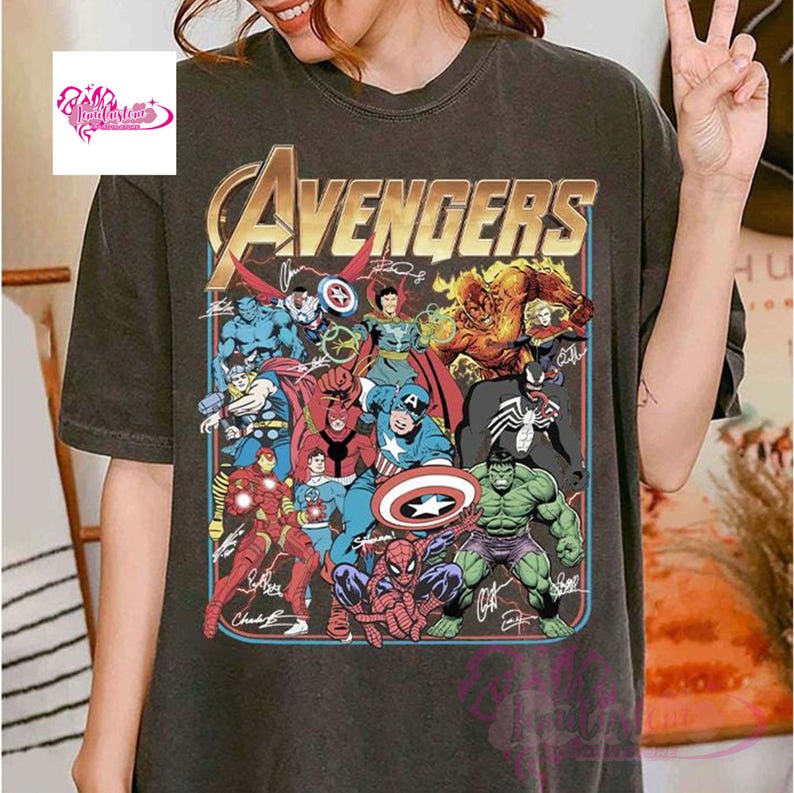 May include: Dark gray t-shirt featuring a colorful graphic of the Avengers. The design includes various superhero characters and the word "Avengers" in gold lettering. Signatures are visible below the characters.