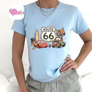 May include: Light blue t-shirt featuring a graphic of Route 66 with the number 66 in a white sign. The graphic includes cartoon cars, a gas pump, and the words "Cozy Cone Motel".