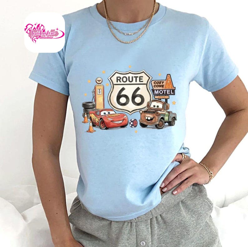 May include: Light blue t-shirt featuring a graphic of Route 66 with the number 66 in a white sign. The graphic includes cartoon cars, a gas pump, and the words "Cozy Cone Motel".