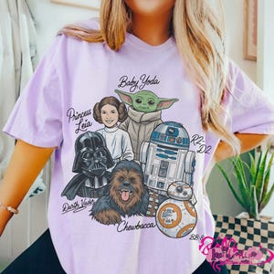 May include: A light purple t-shirt featuring a graphic of Star Wars characters including Baby Yoda, Princess Leia, Darth Vader, Chewbacca, R2-D2, and BB-8. The names of the characters are written in a stylized font. The shirt has short sleeves.