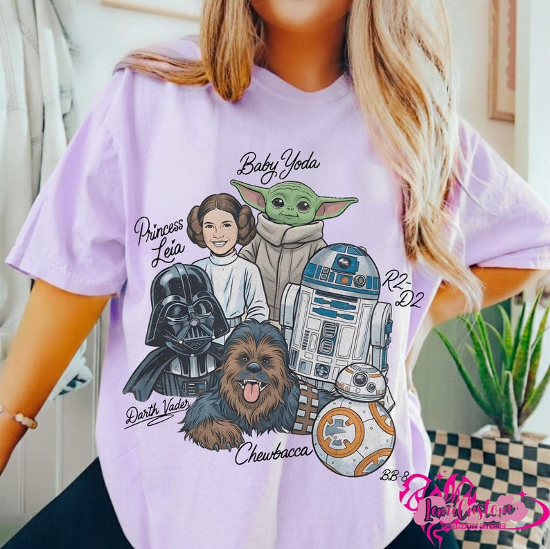 May include: A light purple t-shirt featuring a graphic of Star Wars characters including Baby Yoda, Princess Leia, Darth Vader, Chewbacca, R2-D2, and BB-8. The names of the characters are written in a stylized font. The shirt has short sleeves.