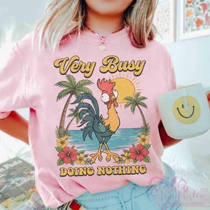 May include: Pale pink t-shirt featuring a cartoon rooster design with palm trees, a sun, and the words "Very Busy Doing Nothing". A white mug with a yellow smiley face is visible in the image.