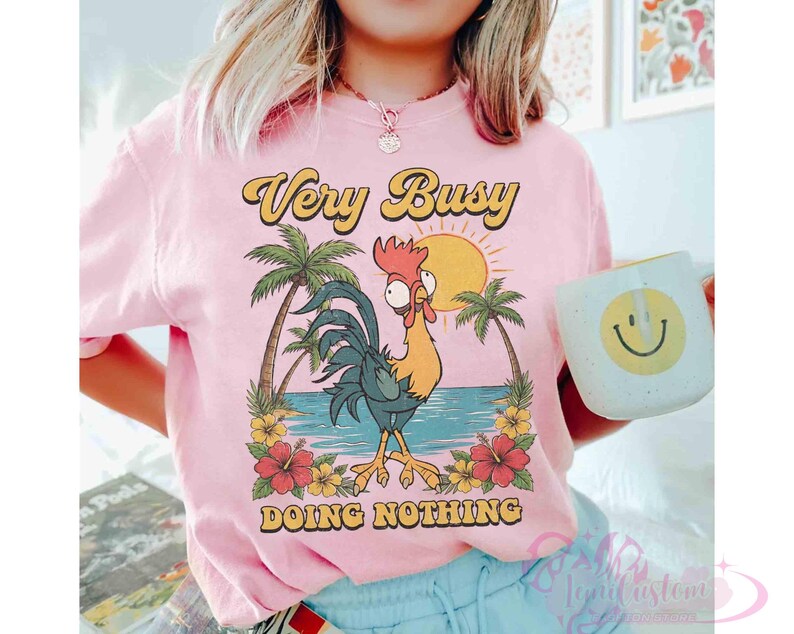 May include: Pale pink t-shirt featuring a cartoon rooster design with palm trees, a sun, and the words "Very Busy Doing Nothing". A white mug with a yellow smiley face is visible in the image.