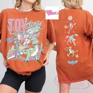 May include: Burnt orange t-shirt featuring the Toy Story logo and characters Woody, Buzz, and Rex. The front of the shirt displays the text "TOY STORY" and "SINCE 1995". The back of the shirt features the characters Woody, Buzz, an alien, and Rex.