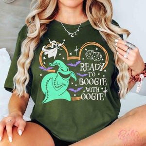 May include: An olive green short-sleeve t-shirt featuring a graphic print of a light green Oogie Boogie character, a white ghost dog, dice, and purple bats. The text reads READY TO BOOGIE WITH OOGIE, arranged within a Mickey Mouse ear outline.