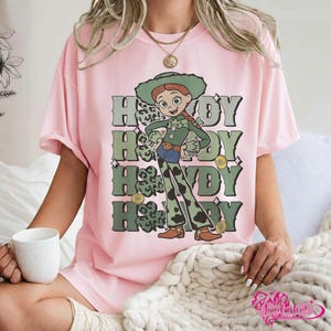May include: Pink t-shirt featuring a cartoon character in a green outfit and hat, with the word "Howdy" repeated four times in green and white. The character has red hair and is wearing brown boots. The shirt is a light pink color.