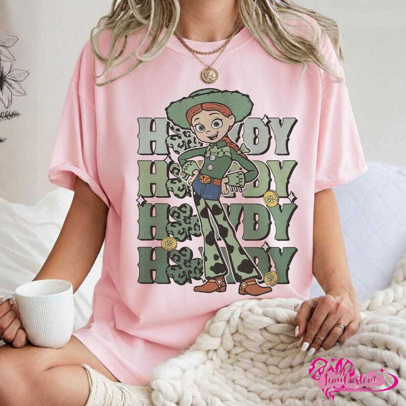 May include: Pink t-shirt featuring a cartoon character in a green outfit and hat, with the word "Howdy" repeated four times in green and white. The character has red hair and is wearing brown boots. The shirt is a light pink color.