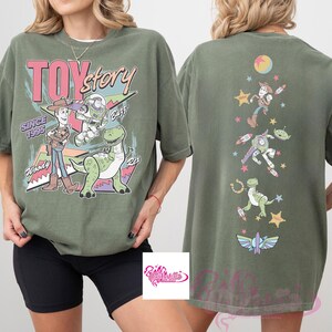 May include: Olive green t-shirt featuring the words "TOY STORY" in pink and white, with Woody, Buzz, and Rex graphics. The back of the shirt has additional character graphics. The text "SINCE 1995" is also visible.