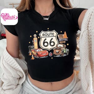 May include: Black crop top featuring a Route 66 graphic with Lightning McQueen and Mater from the Cars movie. The design includes a gas pump, Cozy Cone Motel sign, and stars. The shirt has a round neckline and short sleeves.