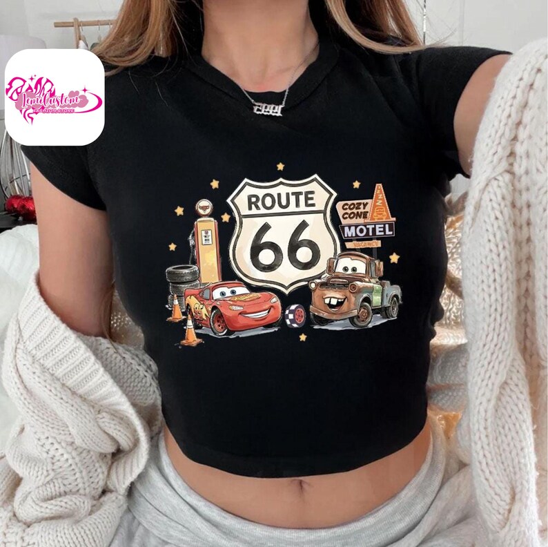 May include: Black crop top featuring a Route 66 graphic with Lightning McQueen and Mater from the Cars movie. The design includes a gas pump, Cozy Cone Motel sign, and stars. The shirt has a round neckline and short sleeves.
