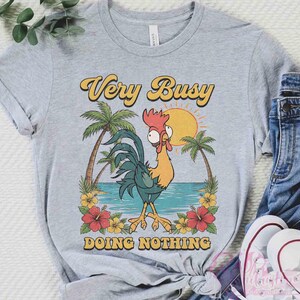 May include: A light grey t-shirt featuring a cartoon rooster design. The text "Very Busy Doing Nothing" is in yellow and orange. The graphic includes palm trees, a sun, and flowers. The shirt is styled with blue jeans and white trainers.